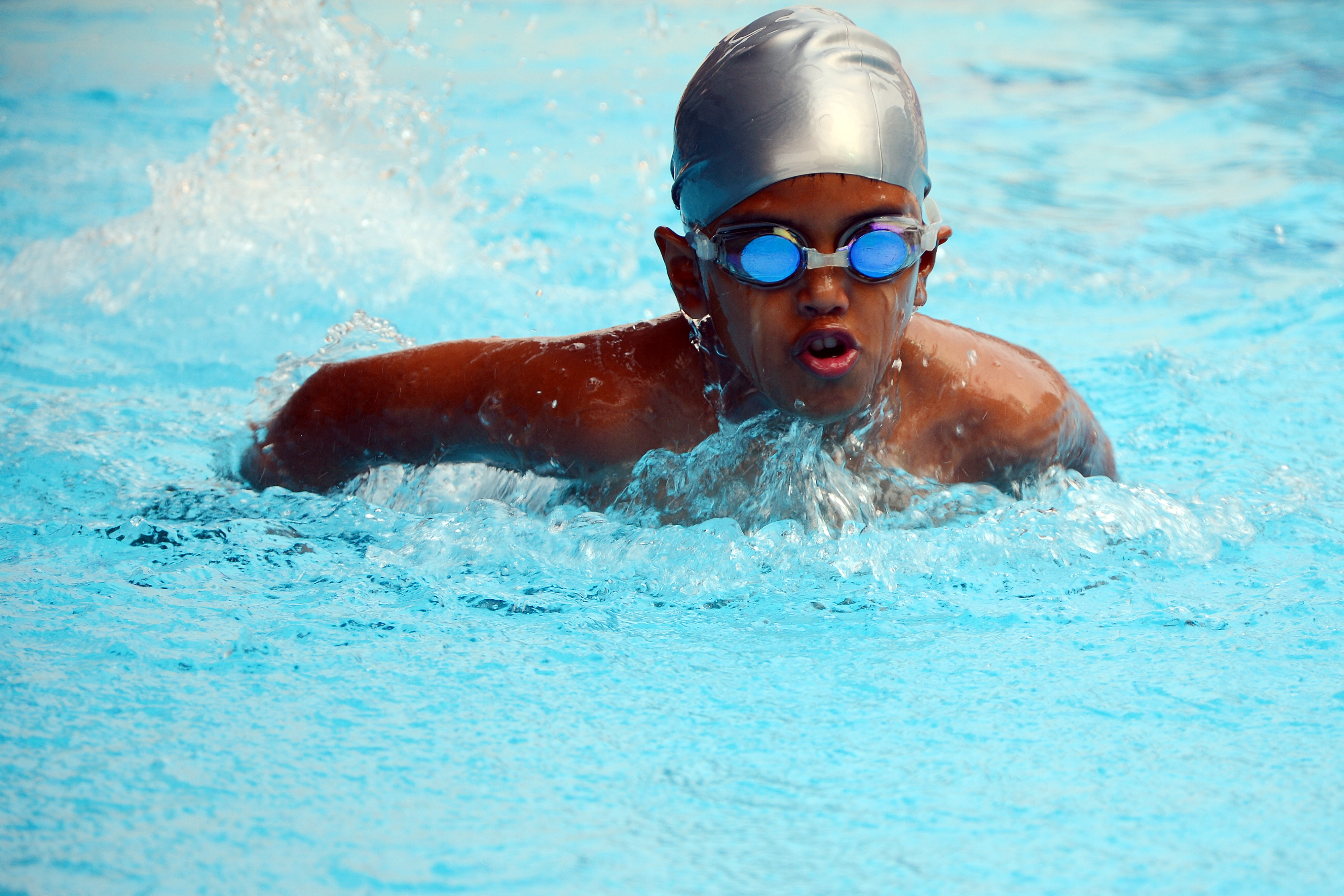 Young boy in sliver swimming cap and blue goggles doing butterfly swim stroke