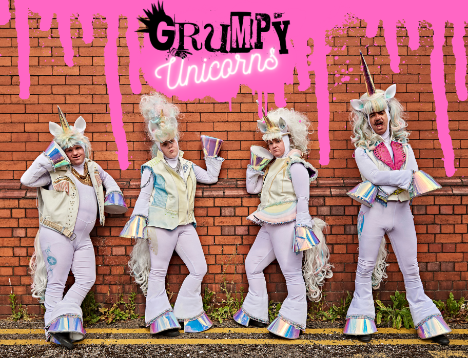 4 adults dressed as unicorns standing against a brick wall.PNG